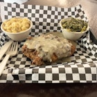 Small bowls of vegan mac and cheese and turnip greens sit on opposite sides of small plastic tray lined with black and white checkered paper. In the center is vegan fried chicken smothered in mushroom sauce. A plastic knife and fork rest on the left-hand side of the tray.